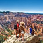 Three hikers with backpacks stand on a cliff edge, overlooking a vast canyon with red rock formations and a clear blue sky, embodying the spirit of a Southern Utah National Parks Tour. three people on a Southern Utah national parks tour, overlooking vista