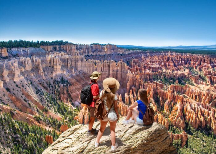 Three hikers with backpacks stand on a cliff edge, overlooking a vast canyon with red rock formations and a clear blue sky, embodying the spirit of a Southern Utah National Parks Tour.