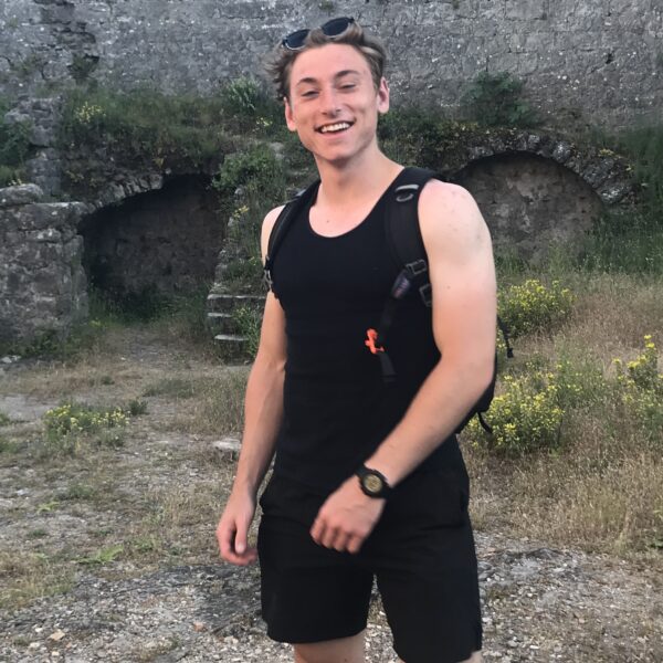 Aiden Gallic, in a black tank top and shorts, stands outdoors with a radiant smile. Behind him, a stone wall mingles with vibrant plants that capture the essence of the scene.