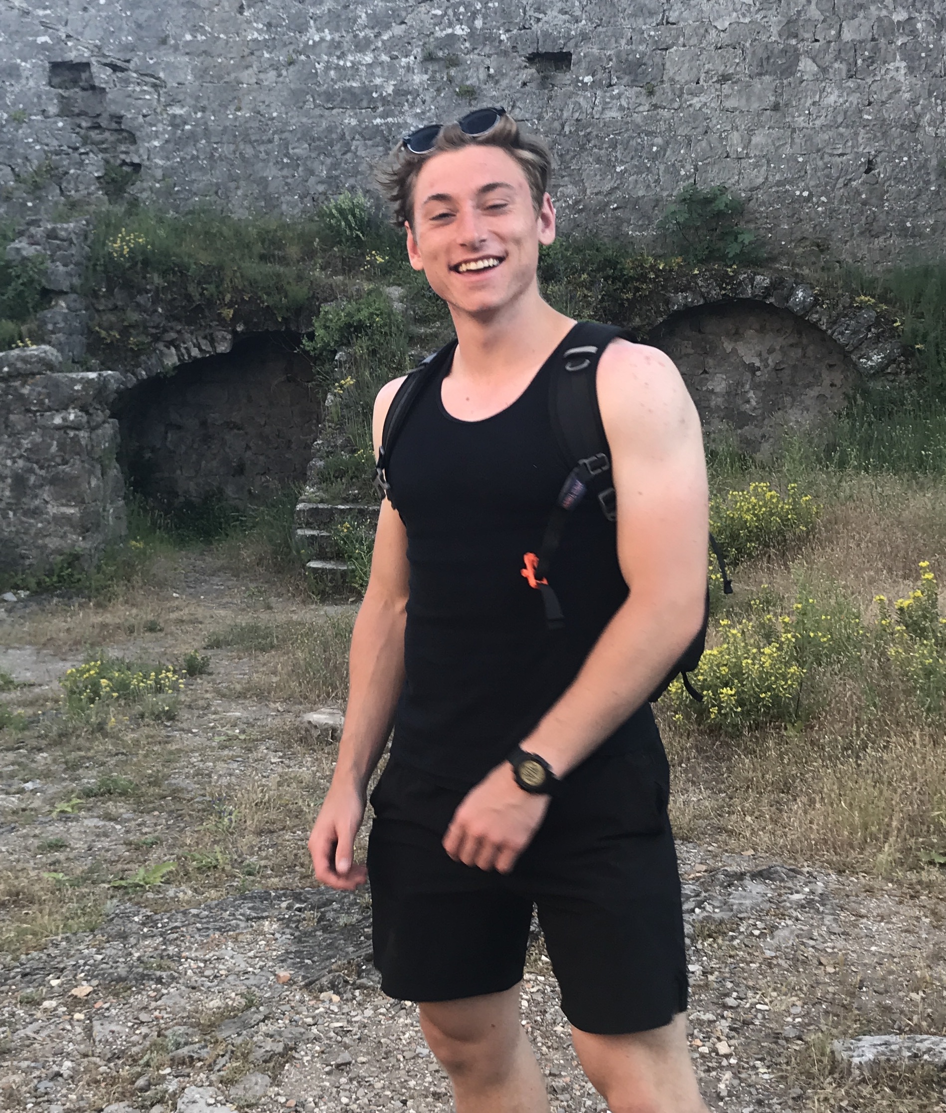 Aiden Gallic, in a black tank top and shorts, stands outdoors with a radiant smile. Behind him, a stone wall mingles with vibrant plants that capture the essence of the scene.