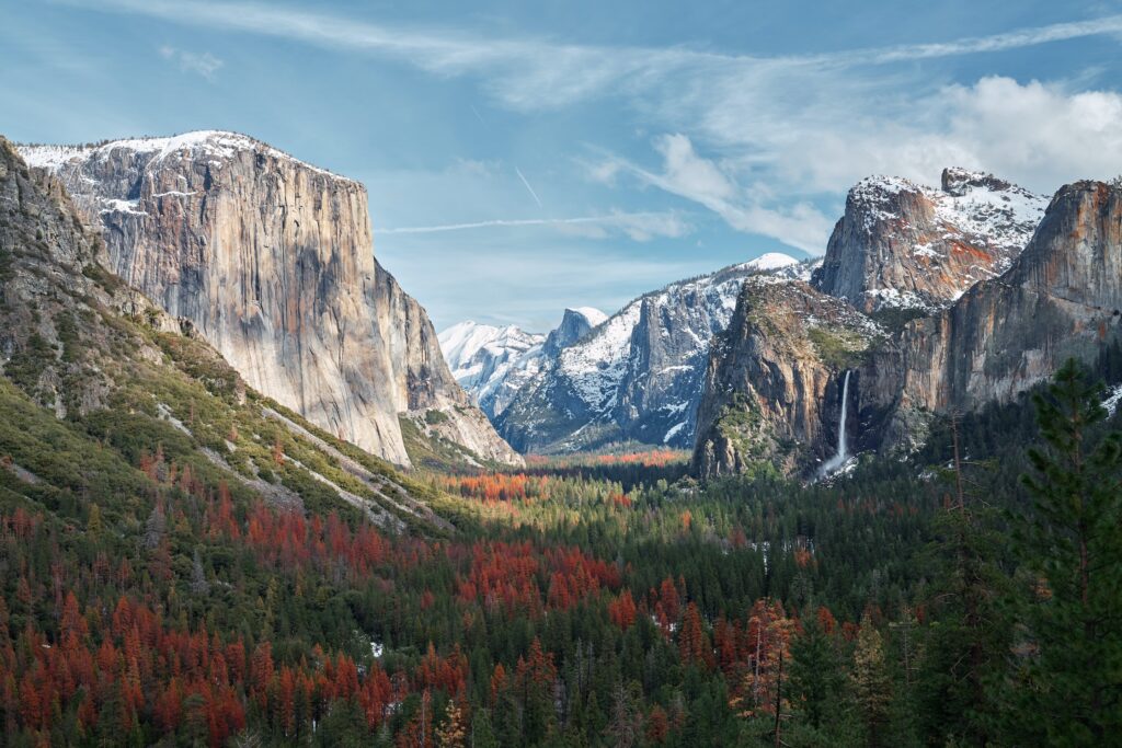 A scenic view of Yosemite Valley with El Capitan on the left, Bridalveil Fall on the right, and Half Dome in the distance, surrounded by forested slopes and dusted with snow