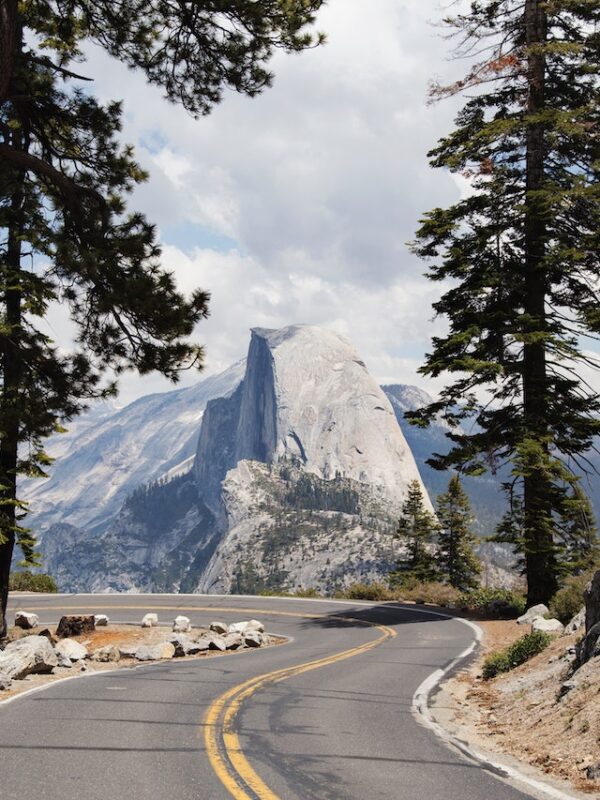 A winding road surrounded by tall trees leads to a view of Half Dome rock formation in Yosemite National Park under partly cloudy skies, perfect for those seeking unforgettable experiences on Yosemite camping tours.