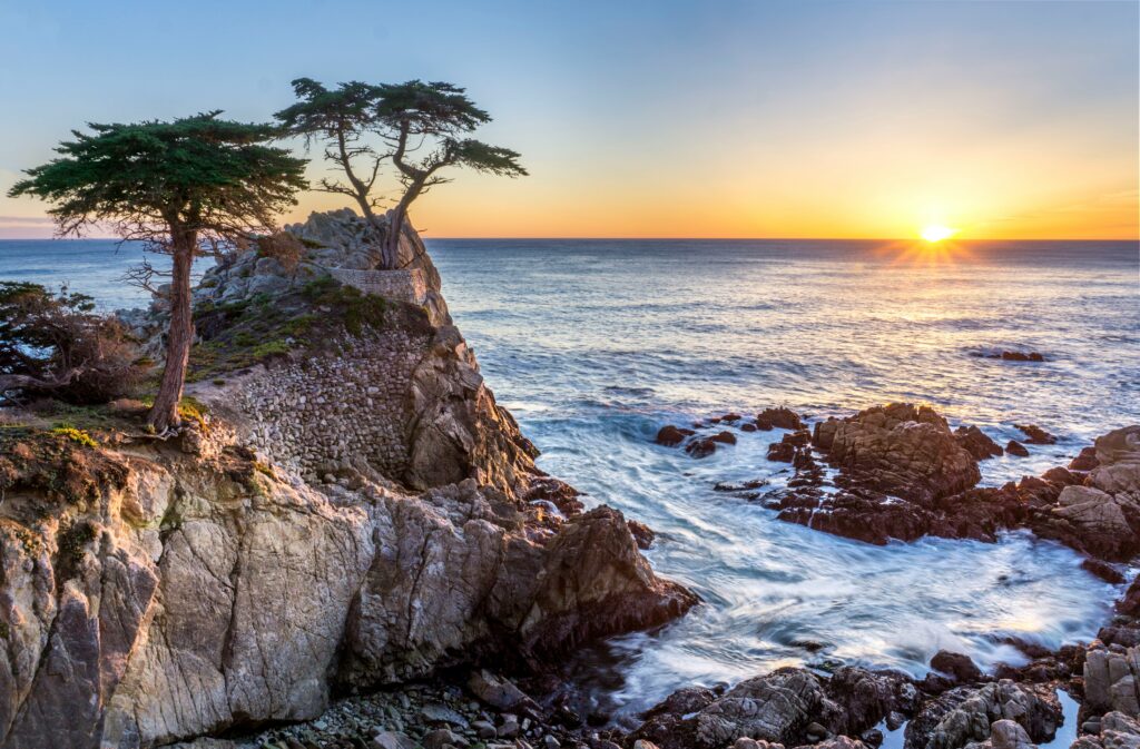 Sunset over a rocky coastline with waves crashing against cliffs and a lone tree atop a promontory during one of the Carmel by the Sea tours.