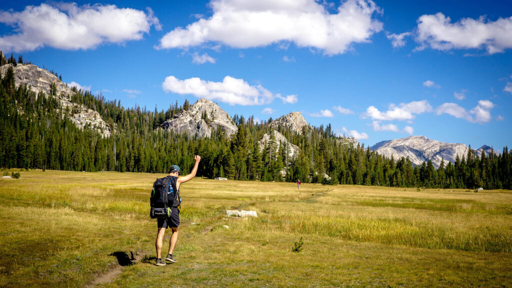 A man in a backpack is walking through a meadow with mountains in the background during a small group tour in California.