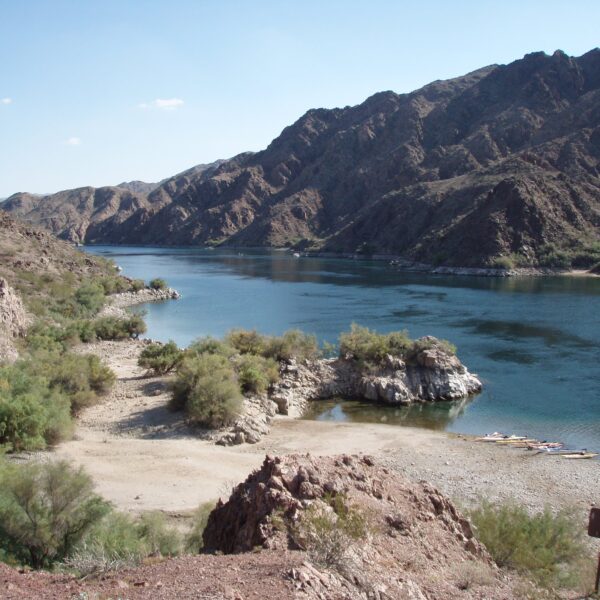 A calm river flows between rocky, brown mountains under a clear sky, with a sandy shoreline and sparse vegetation in the foreground—an ideal setting for a Colorado River kayaking tour near Las Vegas.