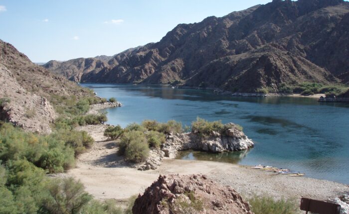 A calm river flows between rocky, brown mountains under a clear sky, with a sandy shoreline and sparse vegetation in the foreground—an ideal setting for a Colorado River kayaking tour near Las Vegas.
