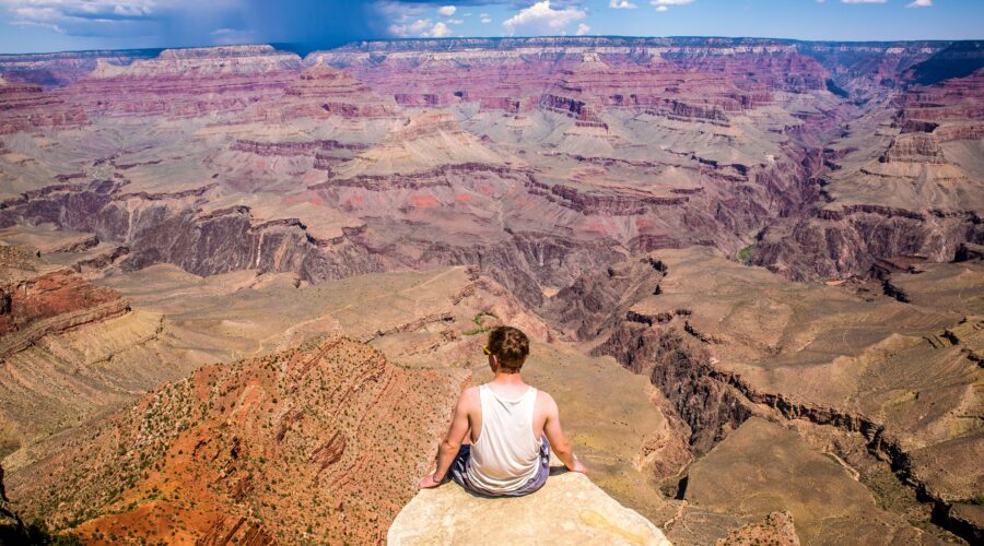 A person sitting on the South Rim, overlooking the vast, colorful landscape of the Grand Canyon under a partly cloudy sky.
