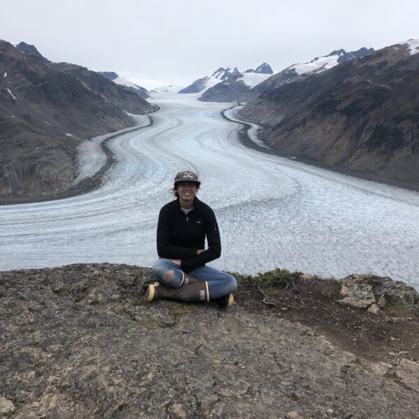 Lana Haugberg sits cross-legged on a rocky ledge, with a majestic glacier and towering mountain range unfolding behind her under the cloudy sky.