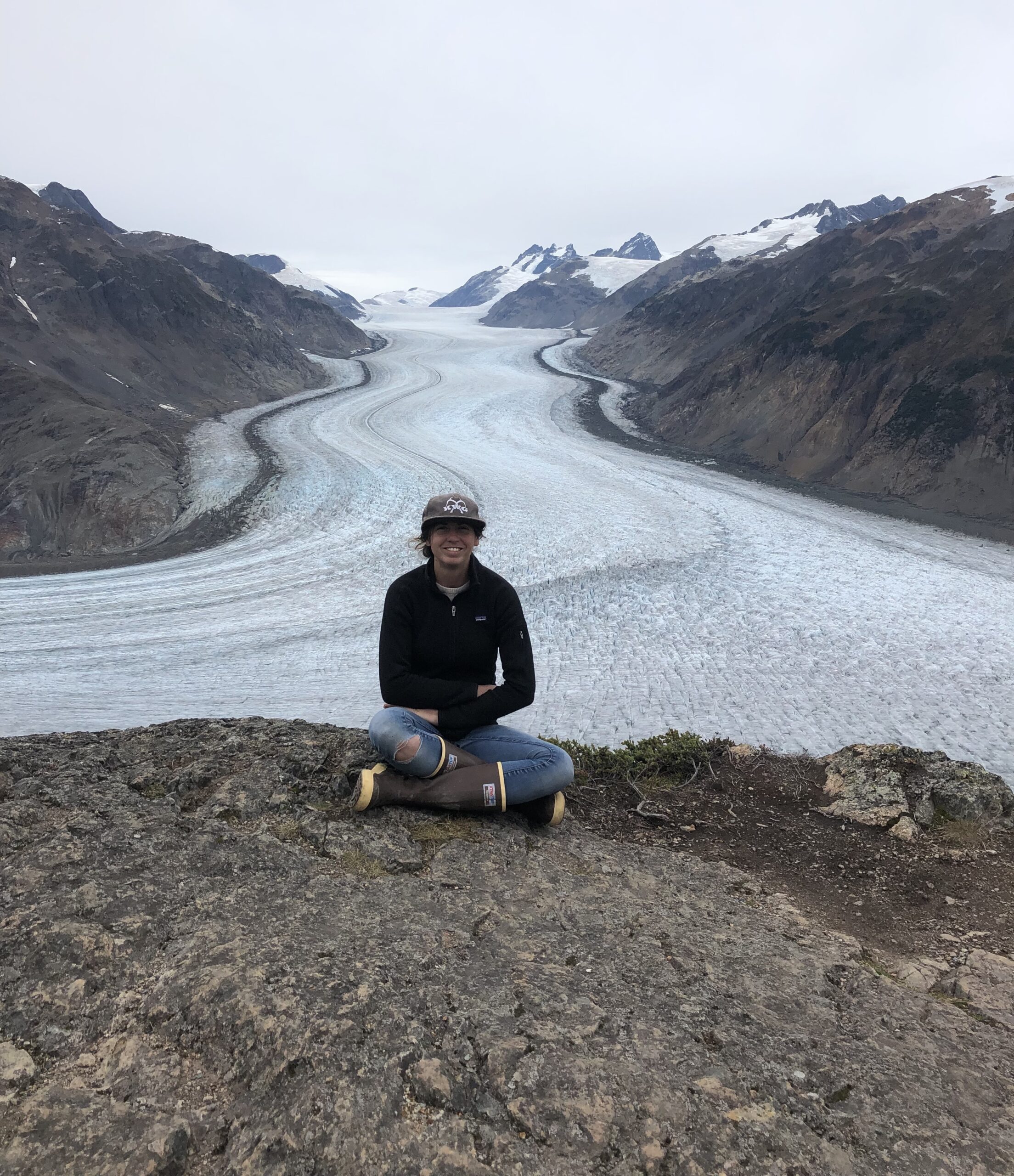 Lana Haugberg sits cross-legged on a rocky ledge, with a majestic glacier and towering mountain range unfolding behind her under the cloudy sky.