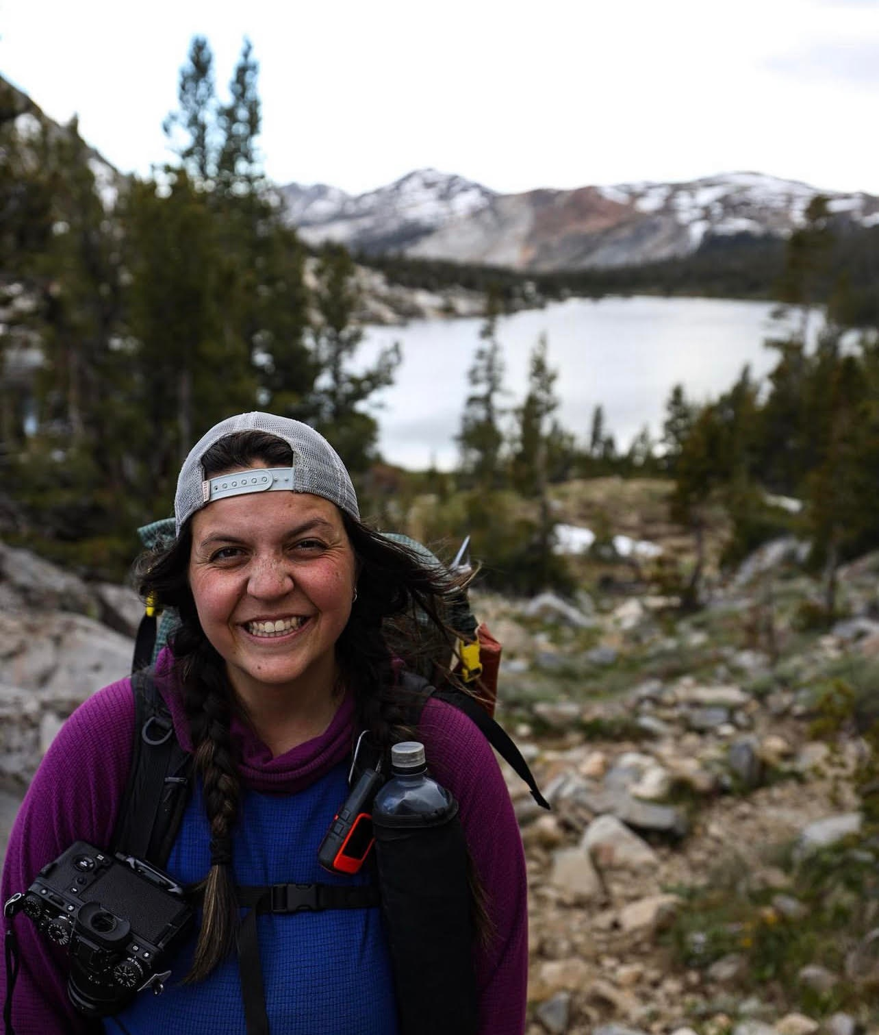 Annie Nobiling, with a radiant smile, holds her camera while standing on a rocky trail. Her hat and backpack hint at adventure, as the trees and lake form a stunning backdrop.