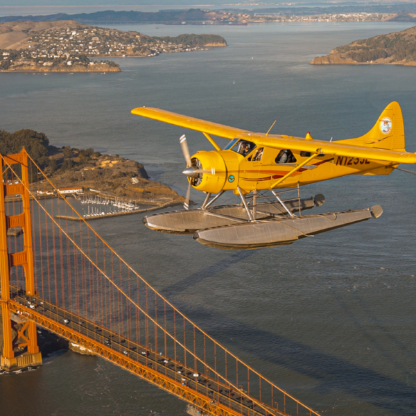 A yellow seaplane from San Francisco seaplane tours flies over the Golden Gate Bridge with the San Francisco Bay and surrounding landscape in the background.