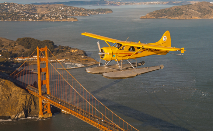 A yellow seaplane from San Francisco seaplane tours flies over the Golden Gate Bridge with the San Francisco Bay and surrounding landscape in the background.