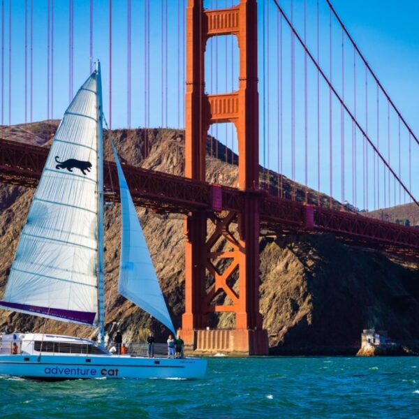 A sailboat named "adventure cat" sails under the golden gate bridge on a sunny day during a San Francisco Bay sailing tour.