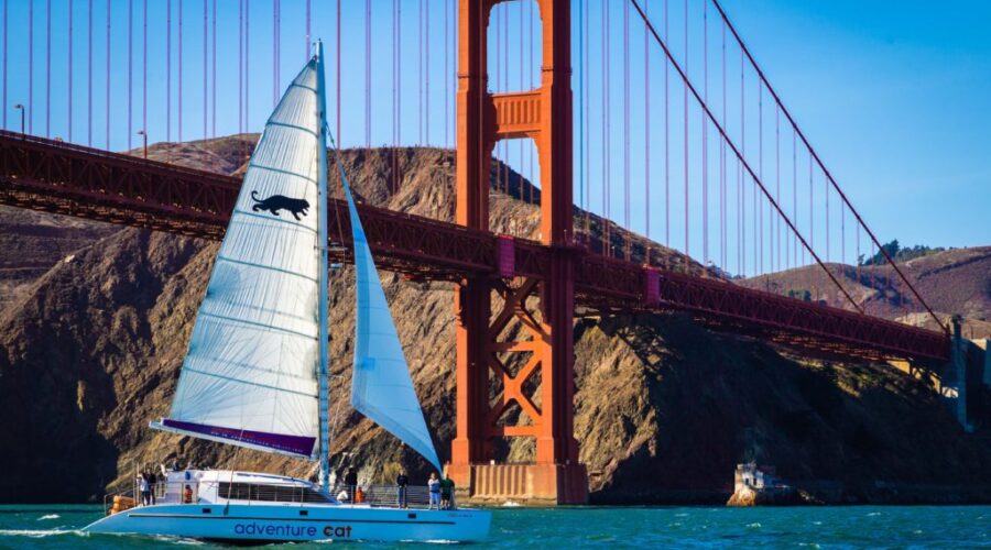 A sailboat named "adventure cat" sails under the golden gate bridge on a sunny day during a San Francisco Bay sailing tour.