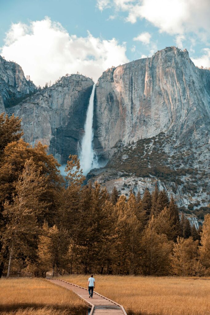 A solitary non-hiker walks towards a towering waterfall cascading down a cliff amidst the forested landscape of Yosemite National Park.