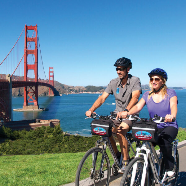 Two people wearing helmets and sunglasses ride bicycles on a path near the Golden Gate Bridge, with clear blue sky and water in the background, perhaps enjoying a day that includes a Sausalito tour or an adventure through Muir Woods.