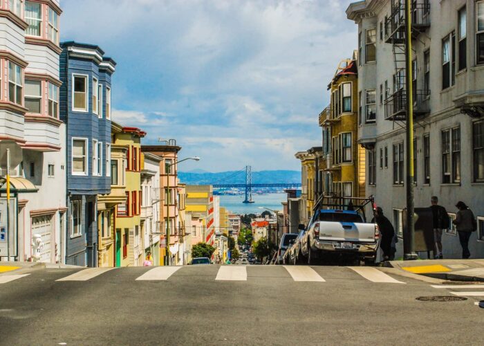 A street view from San Francisco showing steep, colorful residential buildings, pedestrians, and distant views of the bay and a bridge.