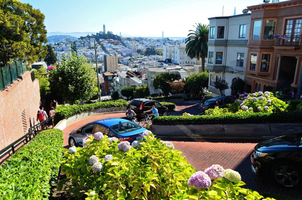 A winding, steep street lined with flowers and greenery, with cars navigating hairpin turns and colorful houses on either side, offers a view of a distant cityscape with blue sky.