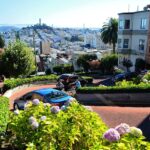 A winding, steep street lined with flowers and greenery, with cars navigating hairpin turns and colorful houses on either side, offers a view of a distant cityscape with blue sky.