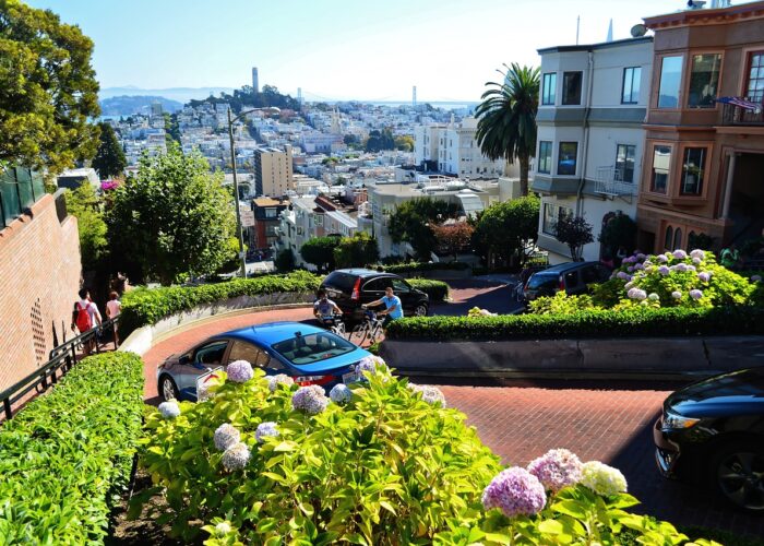 A winding, steep street lined with flowers and greenery, with cars navigating hairpin turns and colorful houses on either side, offers a view of a distant cityscape with blue sky.