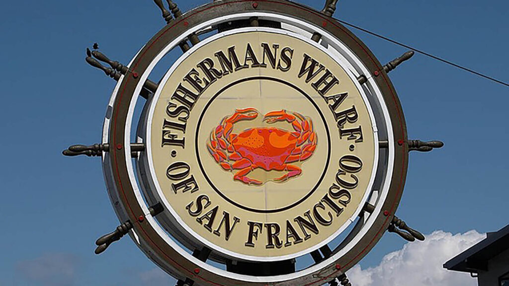 A large sign with a red crab inside a ship's wheel reads "Fisherman's Wharf of San Francisco" set against a blue sky background.