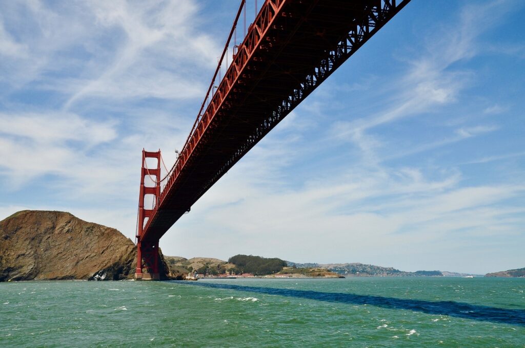 A view of the Golden Gate Bridge from below, showing its red structure spanning across a body of water with hills in the background under a partly cloudy sky.