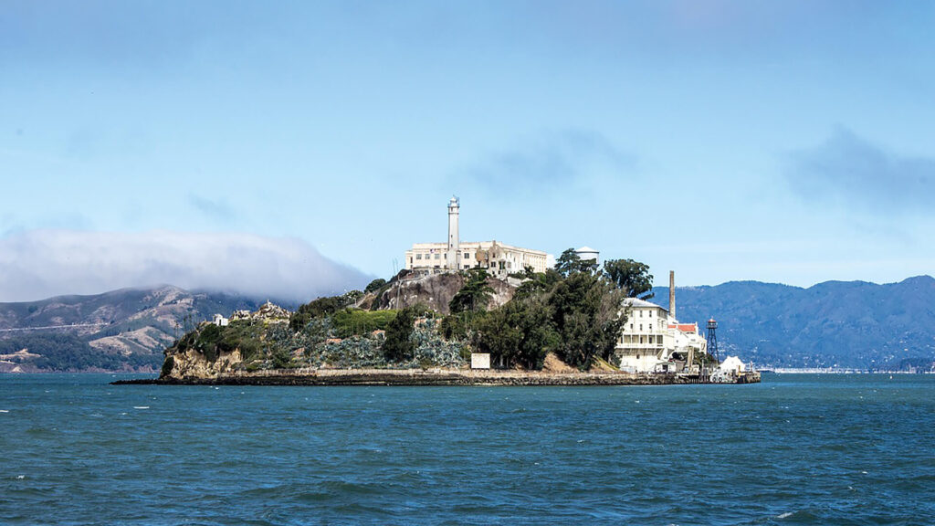 A view of Alcatraz Island in San Francisco Bay, featuring the prison complex and a lighthouse, surrounded by water with distant hills partially covered by clouds.