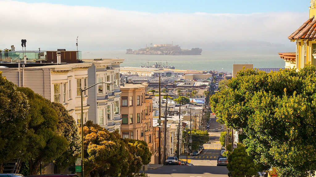 A street in San Francisco slopes down towards a view of Alcatraz Island in the distance, under a partly cloudy sky.