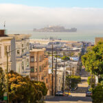 A street in San Francisco slopes down towards a view of Alcatraz Island in the distance, under a partly cloudy sky.