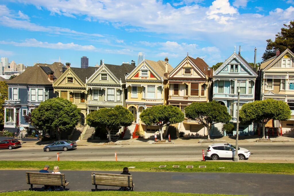 A row of colorful Victorian houses known as the Painted Ladies in San Francisco, with two people sitting on benches in the foreground and cars parked on the street.