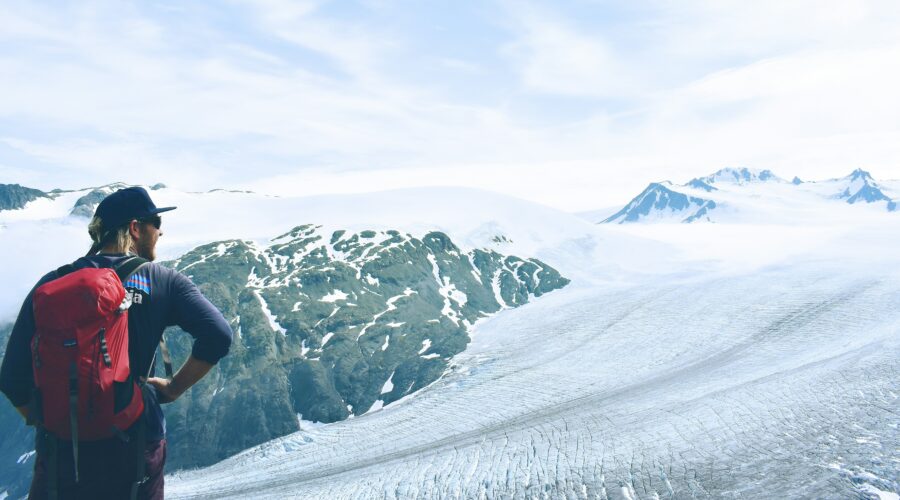 A person with a red backpack stands on a rocky ledge in Alaska, overlooking a vast glacier and snowy mountain range under a partly cloudy sky during their 6-day Highlights Tour.