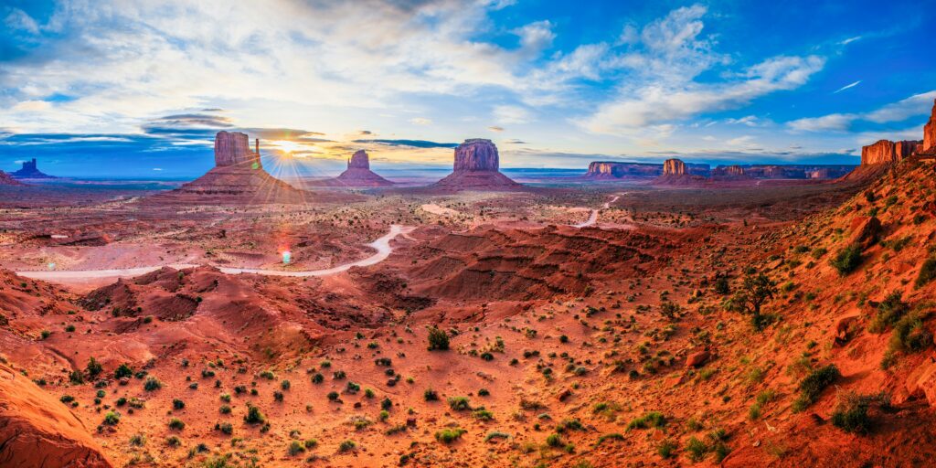 Panoramic view of Monument Valley with sandstone buttes, rocky terrain, and a winding dirt road under a partly cloudy sky during sunrise