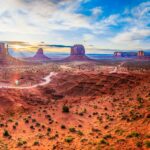 Panoramic view of Monument Valley with sandstone buttes, rocky terrain, and a winding dirt road under a partly cloudy sky during sunrise