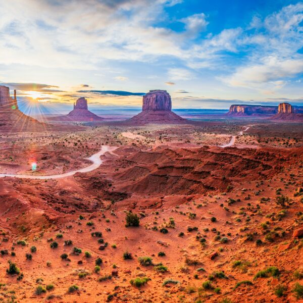 Panoramic view of Monument Valley with sandstone buttes, rocky terrain, and a winding dirt road under a partly cloudy sky during sunrise