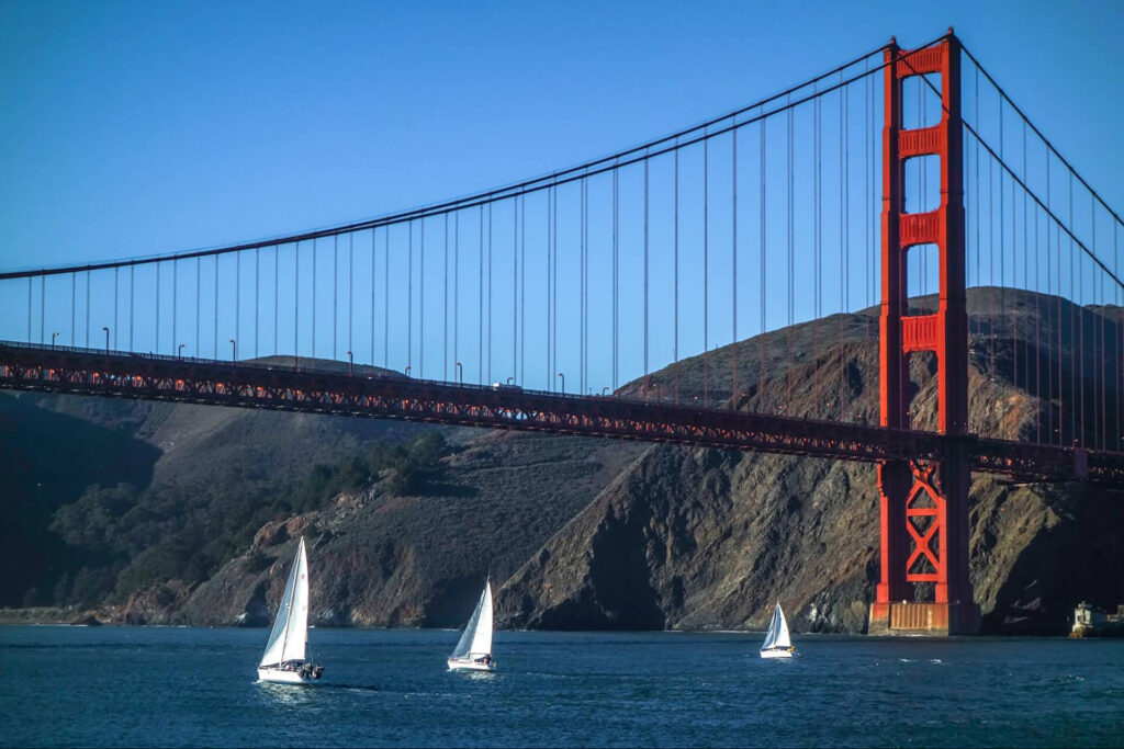 Three sailboats are on the water near the Golden Gate Bridge in San Francisco, with hills in the background and a clear blue sky overhead.