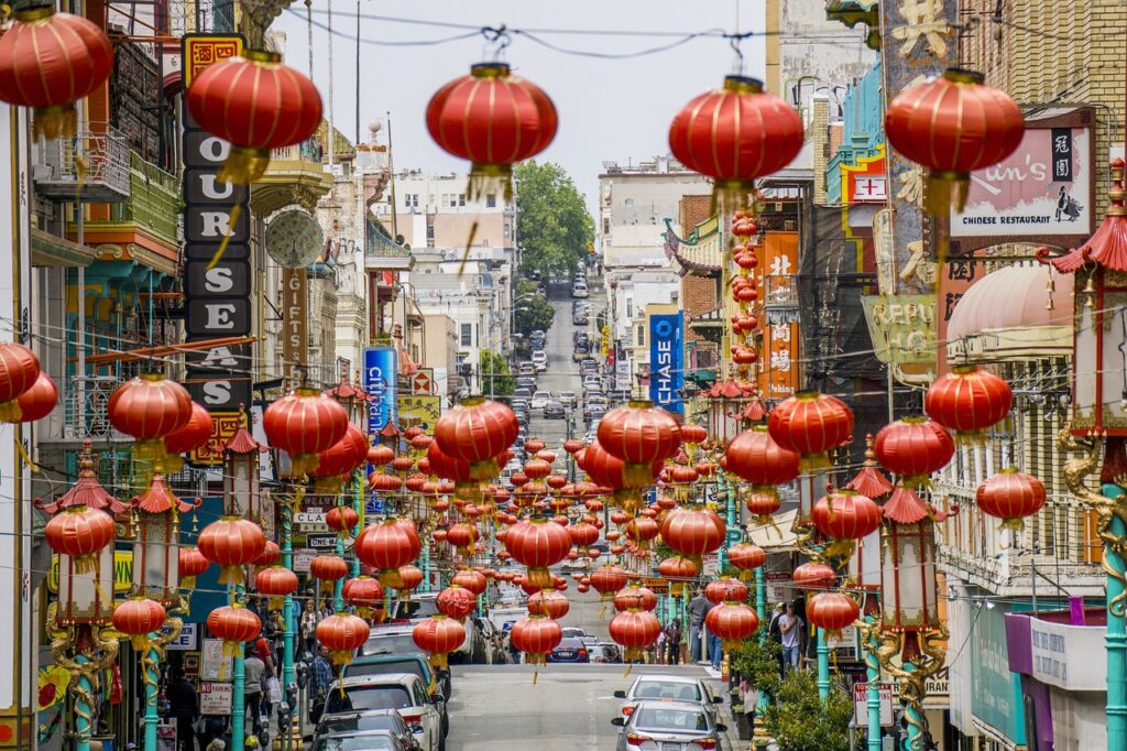 Street decorated with numerous red lanterns hanging above, lined with buildings displaying various signs and advertisements, suggesting a busy urban area in San Francisco likely celebrating a cultural event.