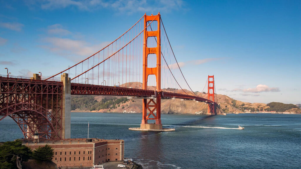 Golden Gate Bridge spans across the water under a clear blue sky, with distant hills in the background and a few boats visible in the bay.