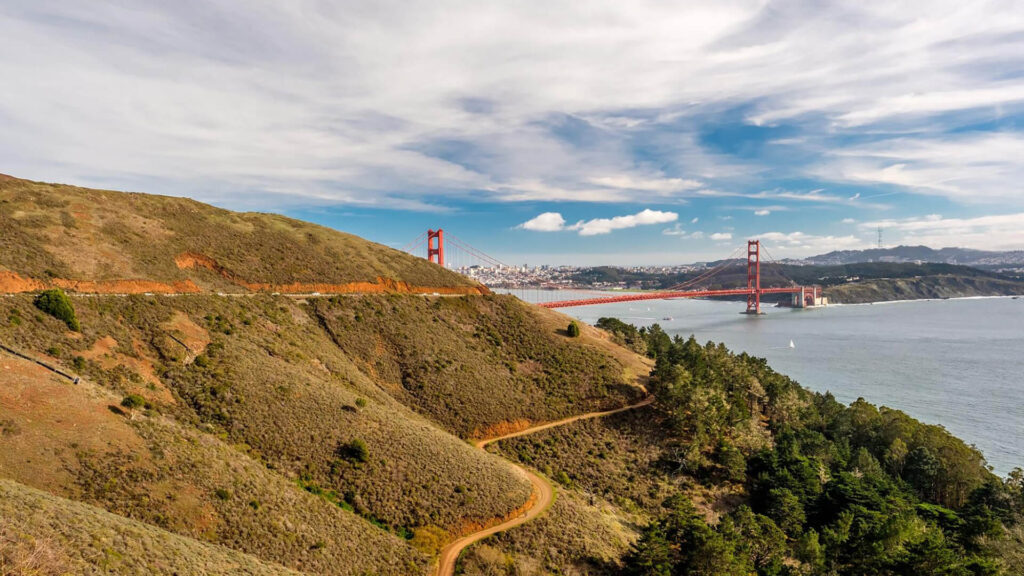 View of the Golden Gate Bridge with surrounding hills, water, and a partly cloudy sky in the background.