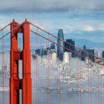A close-up of the Golden Gate Bridge with the San Francisco skyline and mountains in the background, partially obscured by clouds, captures one of the most iconic US cities in all its scenic splendor.