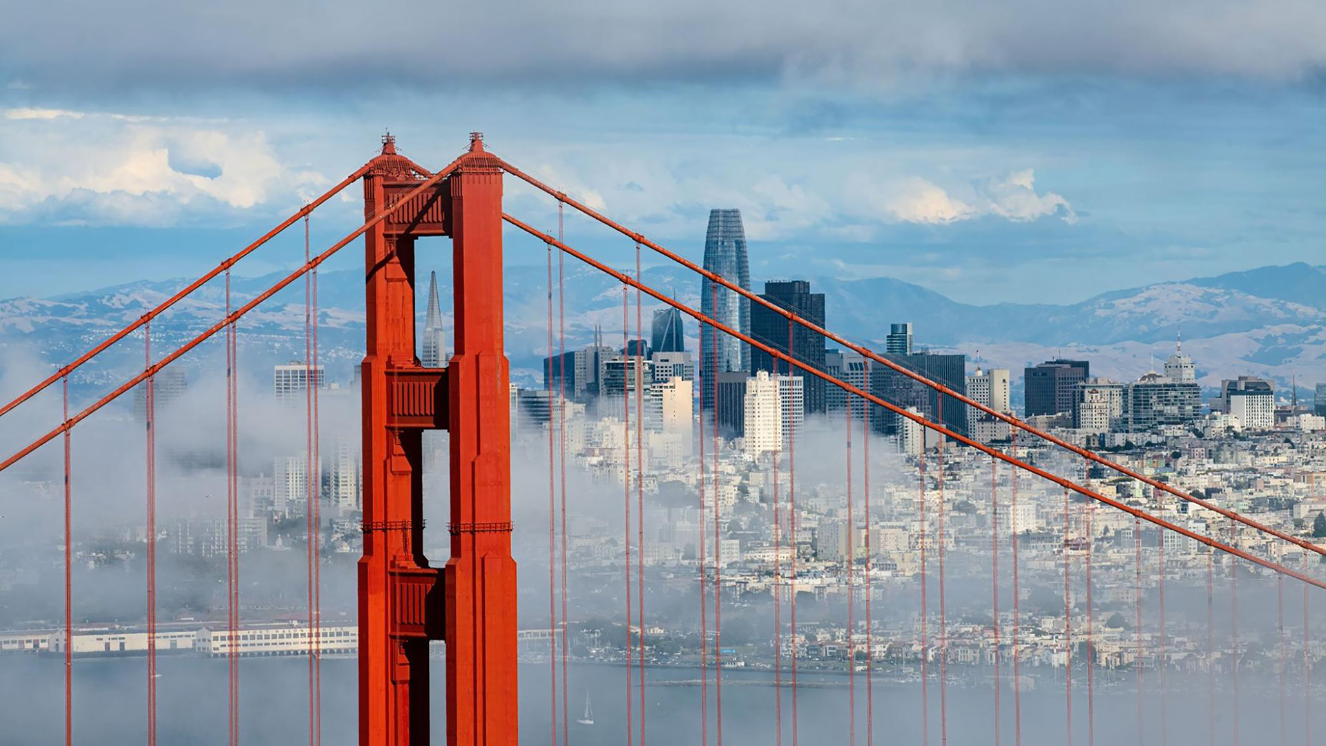 A close-up of the Golden Gate Bridge with the San Francisco skyline and mountains in the background, partially obscured by clouds, captures one of the most iconic US cities in all its scenic splendor.