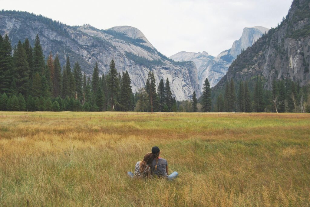 Two people sit on the grass in a meadow, facing away with mountains, trees, and a cloudy sky in the background. It's a serene moment during their Yosemite escape lodging tour.