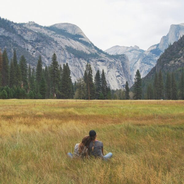 Two people sit on the grass in a meadow, facing away with mountains, trees, and a cloudy sky in the background. It's a serene moment during their Yosemite escape lodging tour.