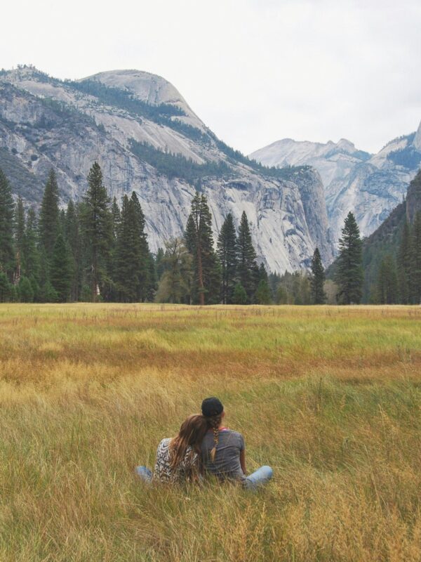 Two people sit on the grass in a meadow, facing away with mountains, trees, and a cloudy sky in the background. It's a serene moment during their Yosemite escape lodging tour.