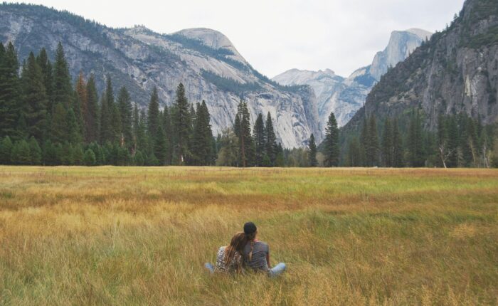 Two people sit on the grass in a meadow, facing away with mountains, trees, and a cloudy sky in the background. It's a serene moment during their Yosemite escape lodging tour.