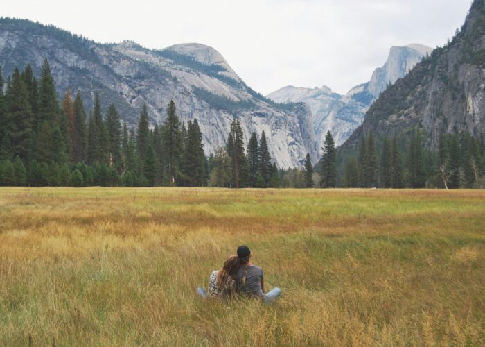 Two people sit on the grass in a meadow, facing away with mountains, trees, and a cloudy sky in the background. It's a serene moment during their Yosemite escape lodging tour.