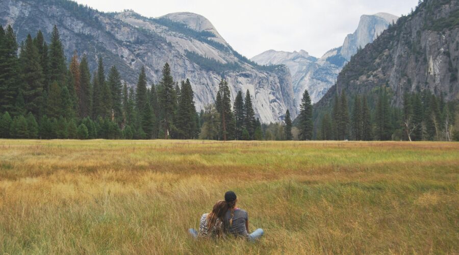 Two people sit on the grass in a meadow, facing away with mountains, trees, and a cloudy sky in the background. It's a serene moment during their Yosemite escape lodging tour.