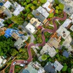 Aerial view of a winding road in San Francisco, surrounded by densely packed houses and greenery. Several cars are driving on the road, and buildings of various shapes and sizes are visible, typical of large US cities.