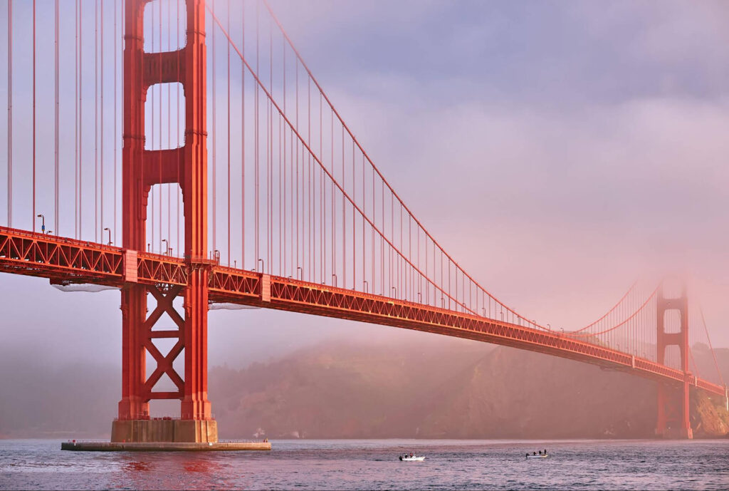 The Golden Gate Bridge stands over a body of water with mist or fog in the background and small boats below.