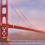 The Golden Gate Bridge stands over a body of water with mist or fog in the background and small boats below.