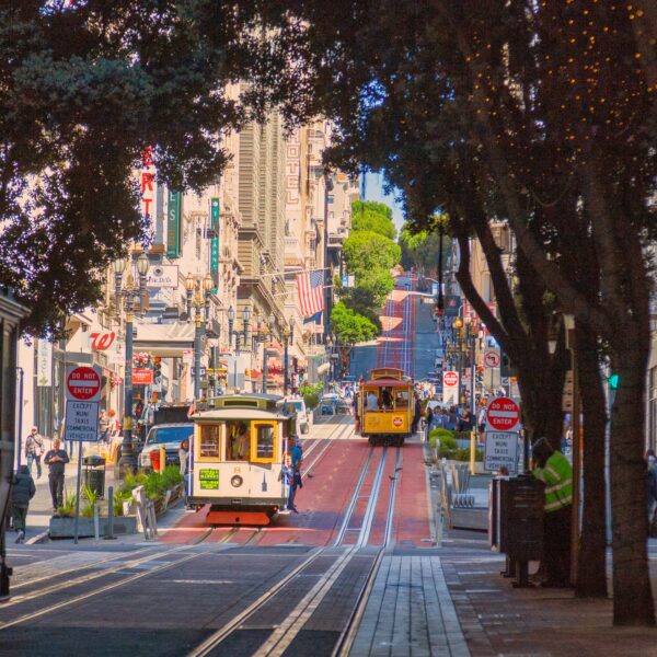 Street view of a city with two cable cars traveling down a hill, surrounded by buildings, trees with string lights, and pedestrians on the sidewalk—a charming scene often featured on the California Parks Highlights Tour from Los Angeles to San Francisco over 5 days.